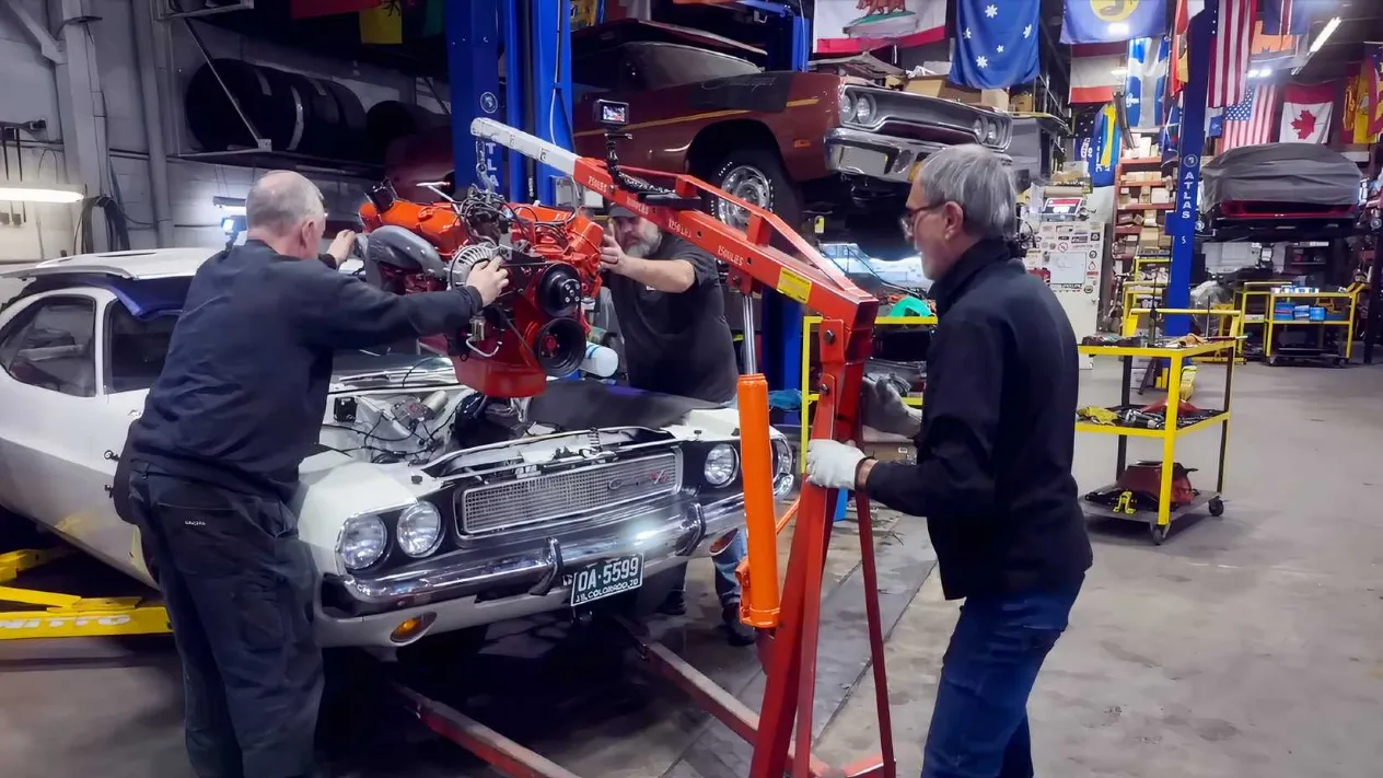 Nick and Robert carefully lowering the original 440 Magnum engine into the Challenger's engine bay using a cherry picker, with tools and garage equipment visible.