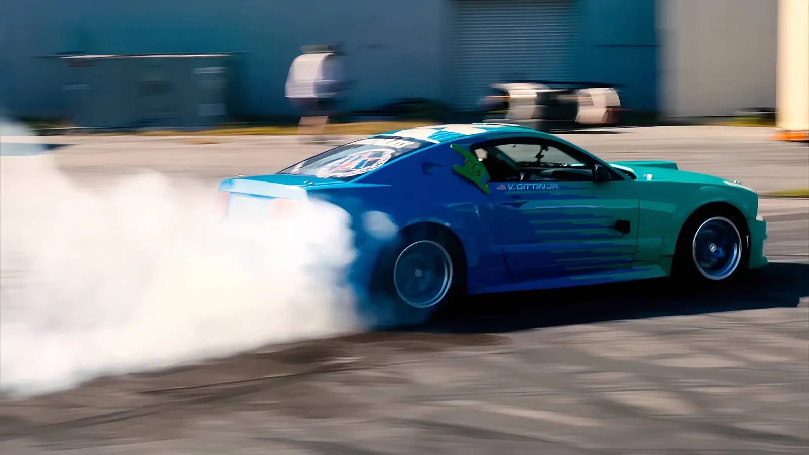 A battle-worn 2005 Mustang drift car sits in a dusty garage, its dark blue paint barely hiding decades of rubber and scars